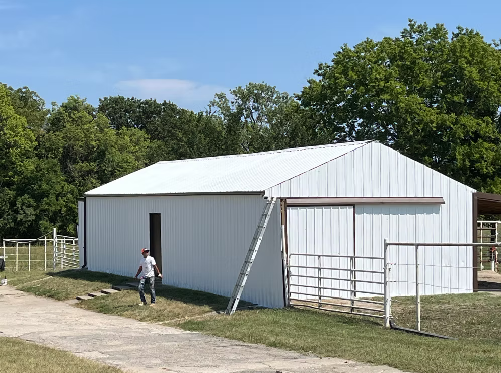 A man standing outside a white metal barn with a ladder leaning against the side, surrounded by grassy and gravel areas and trees in the background on a sunny day.
