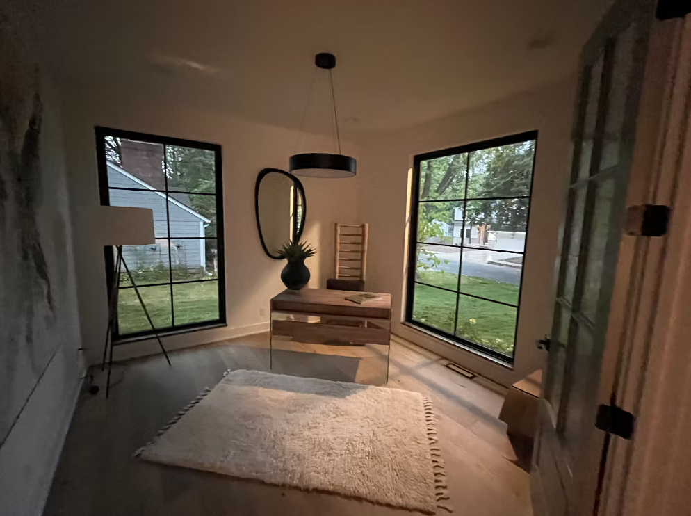 A corner room with two large windows showing a front yard and street outside, a modern desk with a black vase and plant, a tall mirror, a wooden chair, a white textured rug, a floor lamp, and a partially open door.