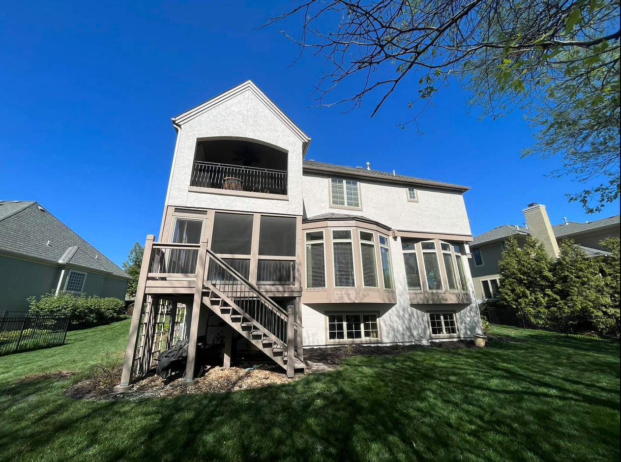 Back view of a modern two-story house with a deck and large windows, surrounded by a well-maintained lawn and neighboring houses, under a clear blue sky.