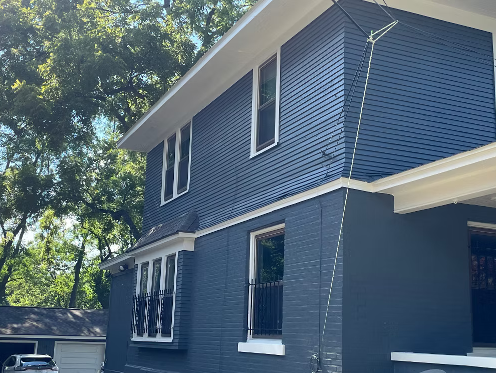 A blue two-story house with white trim and multiple windows, surrounded by greenery, with an outdoor antenna attached to the side of the house.