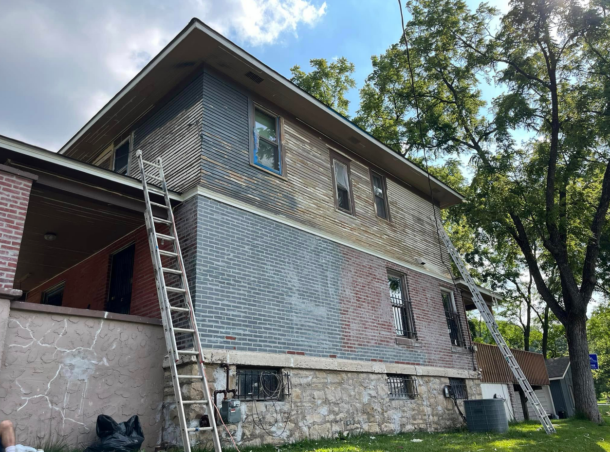 Side view of a two-story house showing the renovation progress. The upper portion has wooden siding, some of which is being replaced. Ladders are leaning against the house, and trees are visible in the background under a partly cloudy sky.