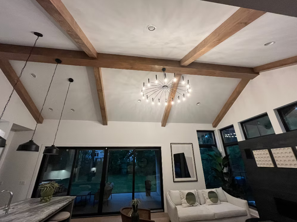 Living room with a high ceiling featuring wooden beams, a modern chandelier, large windows, a white sofa with green pillows, and a sliding glass door leading outside.