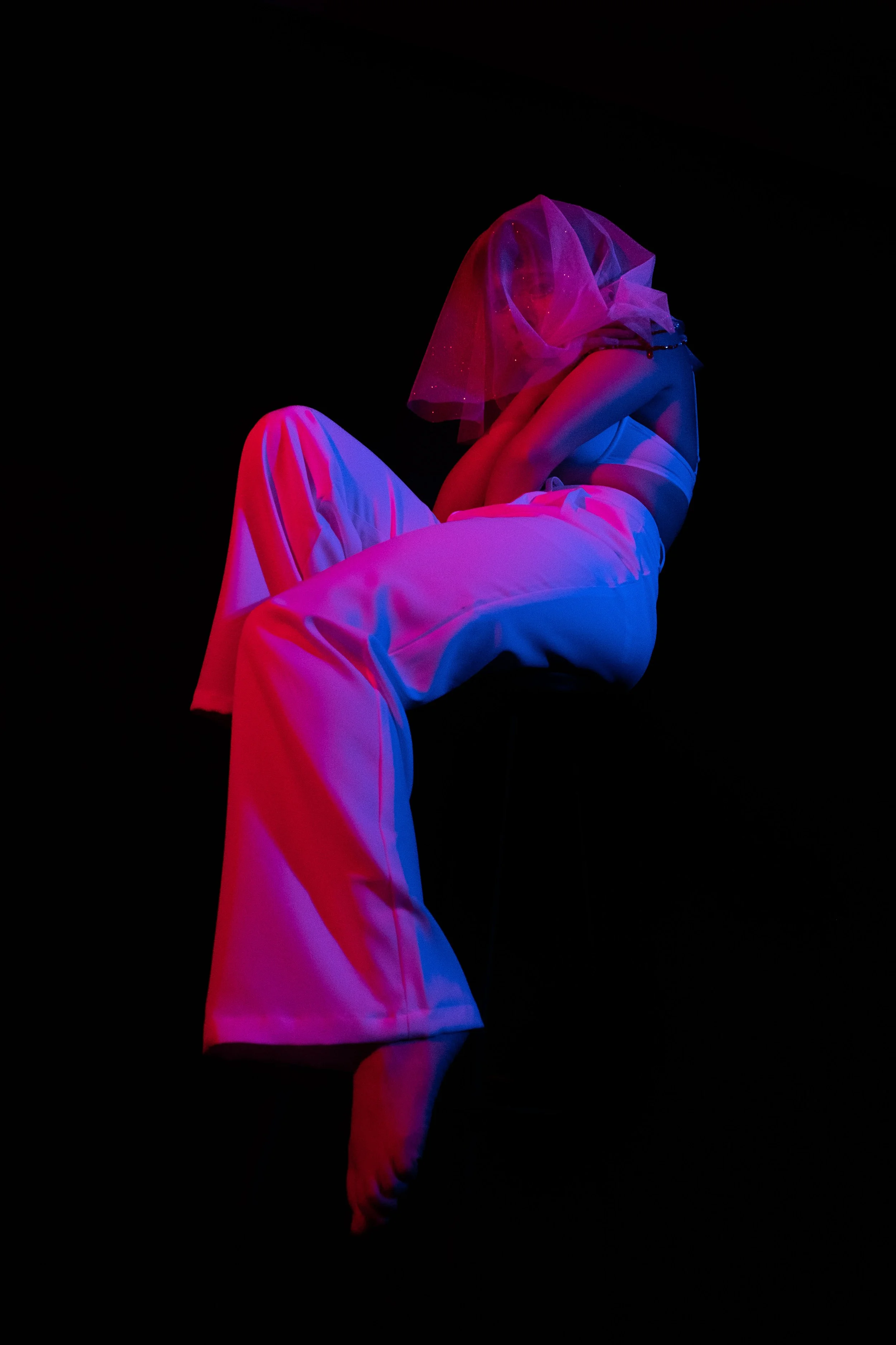 Persona con prenda blanca y velo azul, sentada en posición de cuclillas, en un fondo oscuro, iluminada con luz de neón rosa y azul.