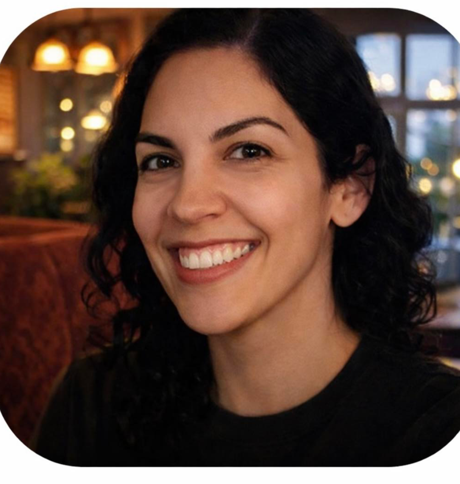 Close-up of a smiling woman with dark curly hair in a warmly lit indoor setting.