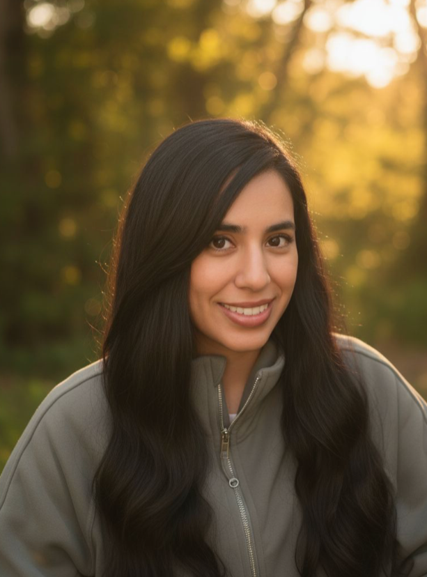 A young woman with long dark hair smiling outdoors during sunset, wearing a gray jacket.