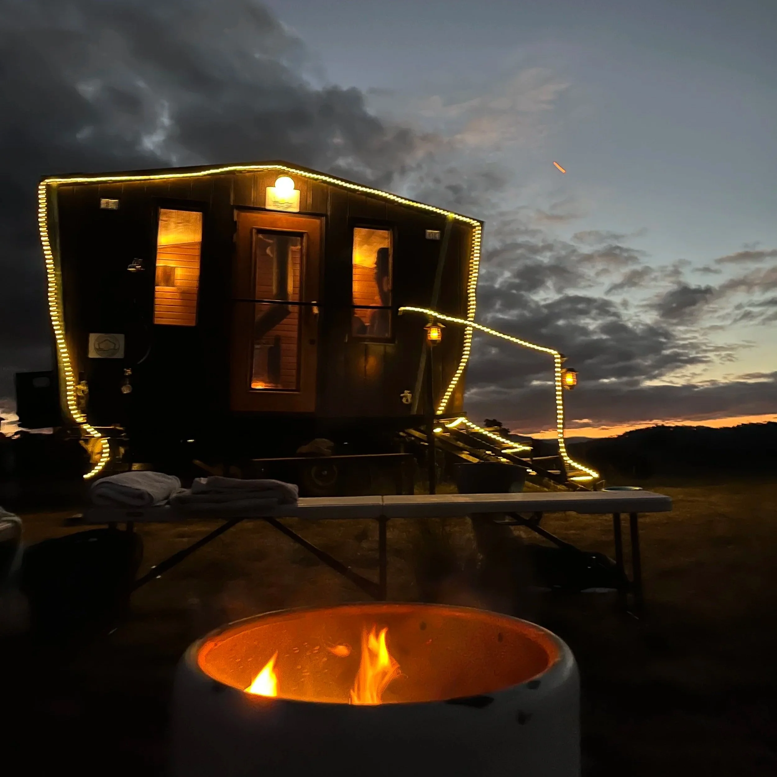 A boat with a wooden structure decorated with yellow lights on a dark evening sky, with a fire pit in the foreground and a cloudy sky overhead.