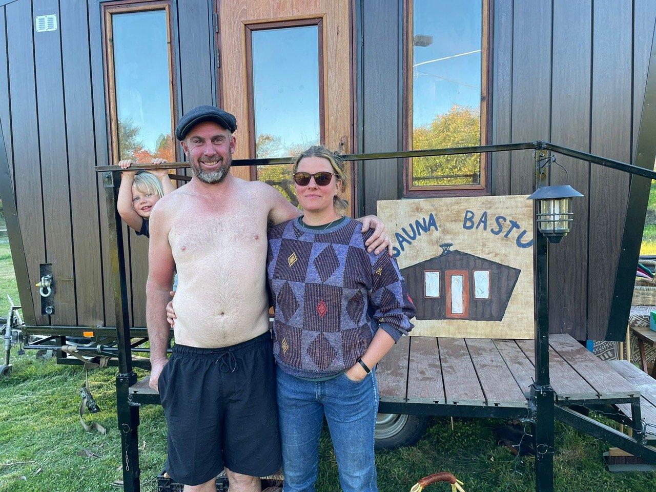 A man with a beard, wearing a beret and black shorts, stands next to a woman with sunglasses and a patterned sweater. A young girl peeks out from behind the man, smiling and holding onto a railing. They are outdoors in front of a small wooden structure with a sign that reads 'Sauna Basto.' The structure has a wooden deck, a lantern light, and window reflections of trees and the sky.