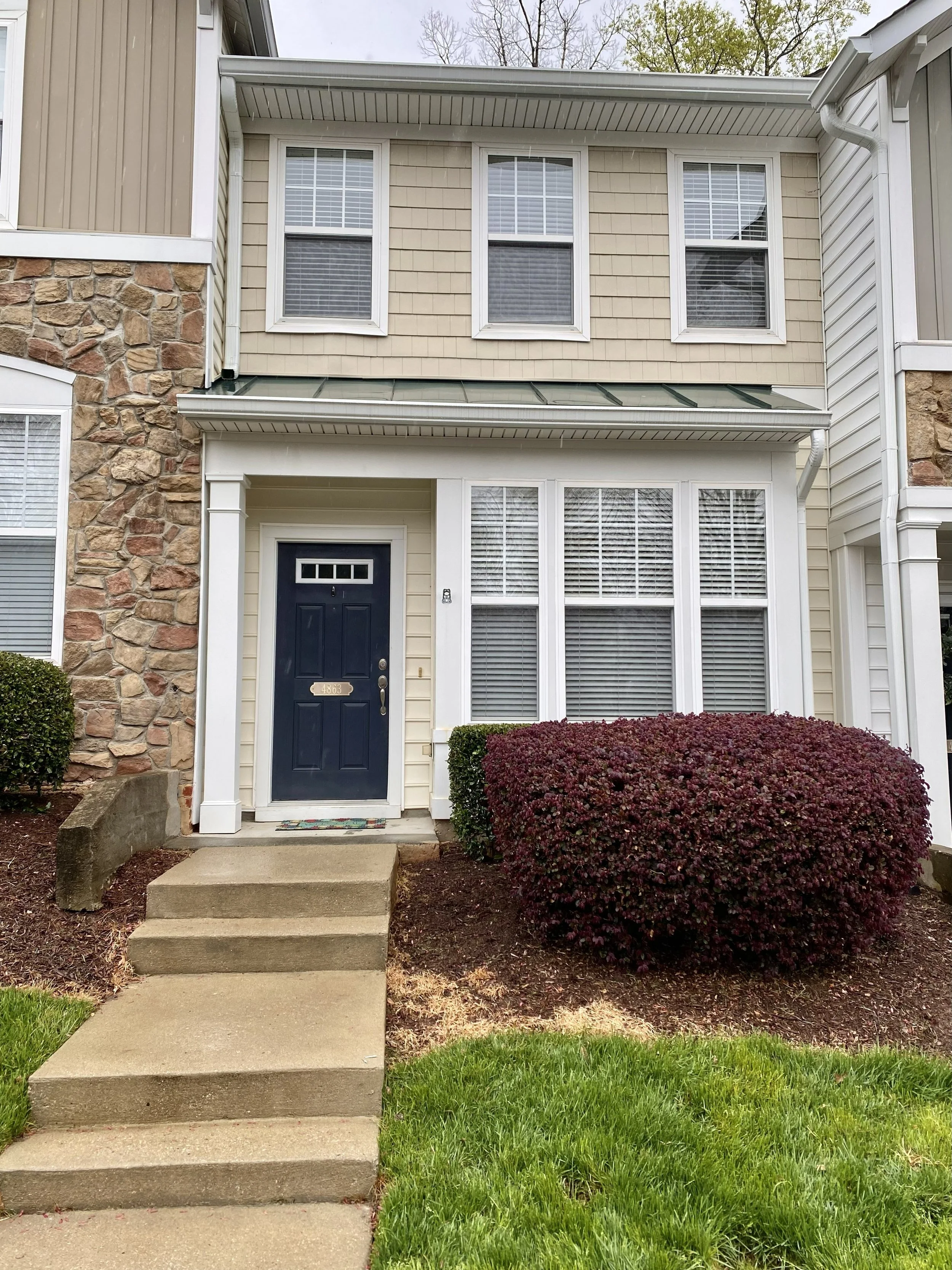 Front view of a two-story house with a navy blue door, beige siding, stone accents, and multiple windows with white blinds. There are steps leading up to the front door, a bush to the right, and a small patch of green grass in the foreground.