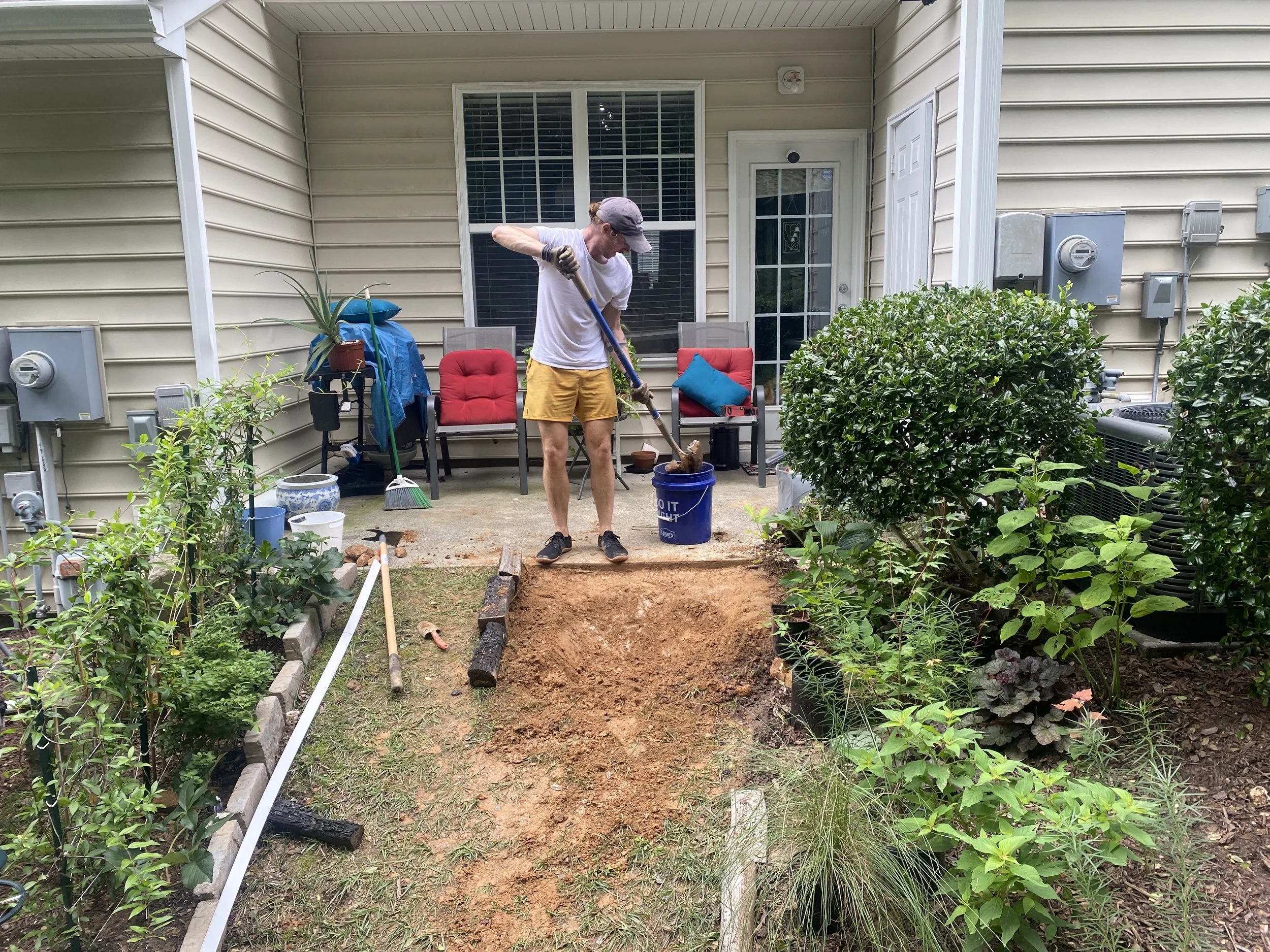 A man with a white shirt and yellow shorts is digging a hole using a shovel. There are two red cushioned chairs, one with a blue pillow, on a patio behind him. Various plants, gardening tools, and potted plants are visible around the area.
