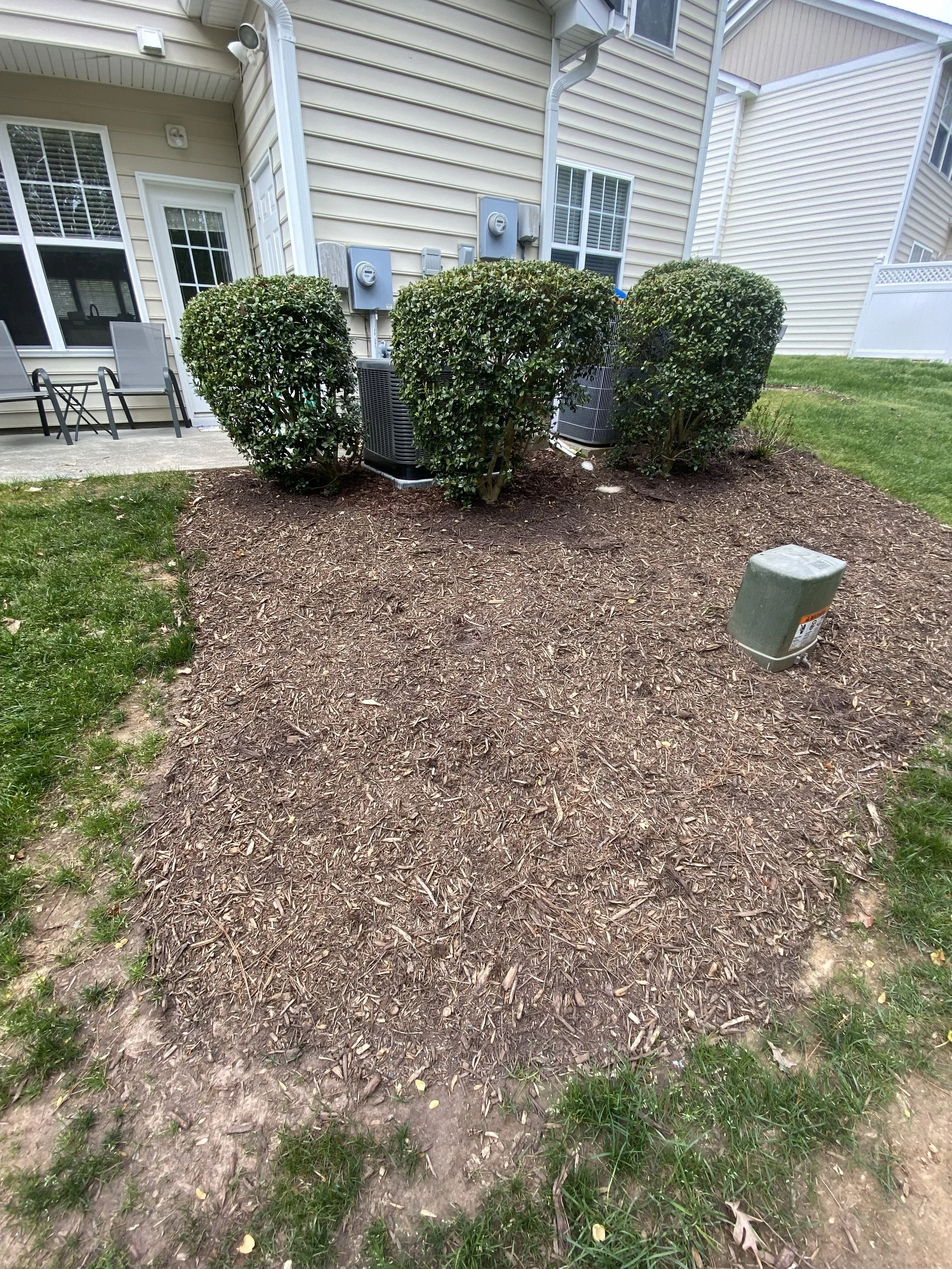 Bare soil with wood mulch in a backyard, surrounded by grass, with a house in the background featuring a door, windows, and bushes near the house HVAC system.