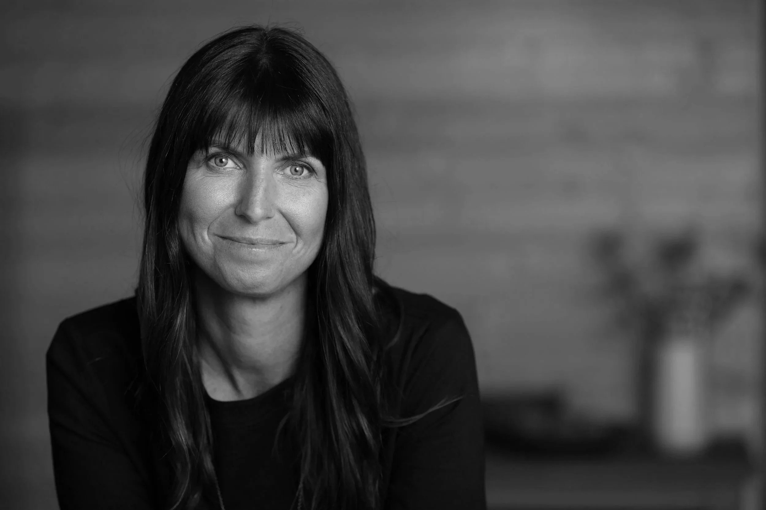A black-and-white portrait photo of a woman with long dark hair and bangs, smiling slightly, wearing a dark top, with a blurred background.