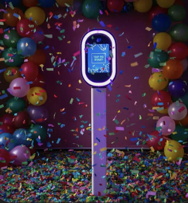 A festive scene with colorful balloons and confetti surrounding a digital kiosk with a bright display, set against a purple background.