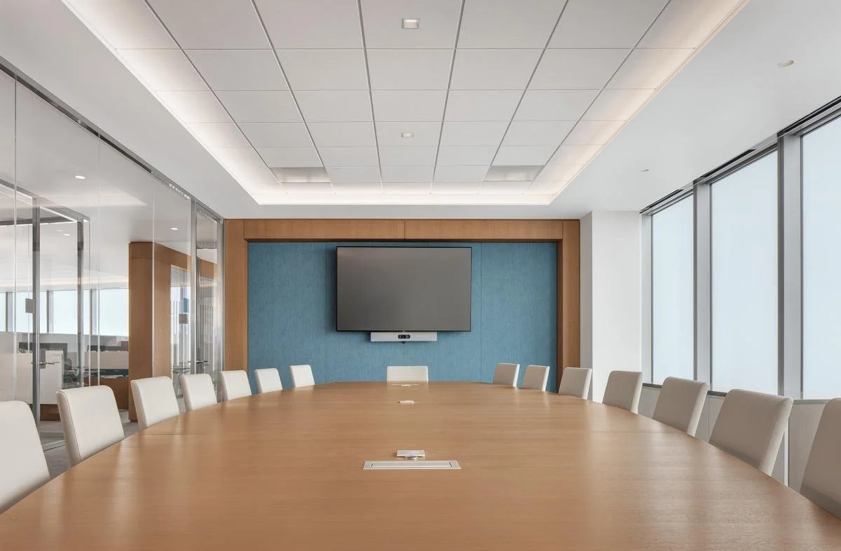 An empty conference room with a long wooden table, white chairs, large windows with blinds, and a wall-mounted television on a blue accent wall.