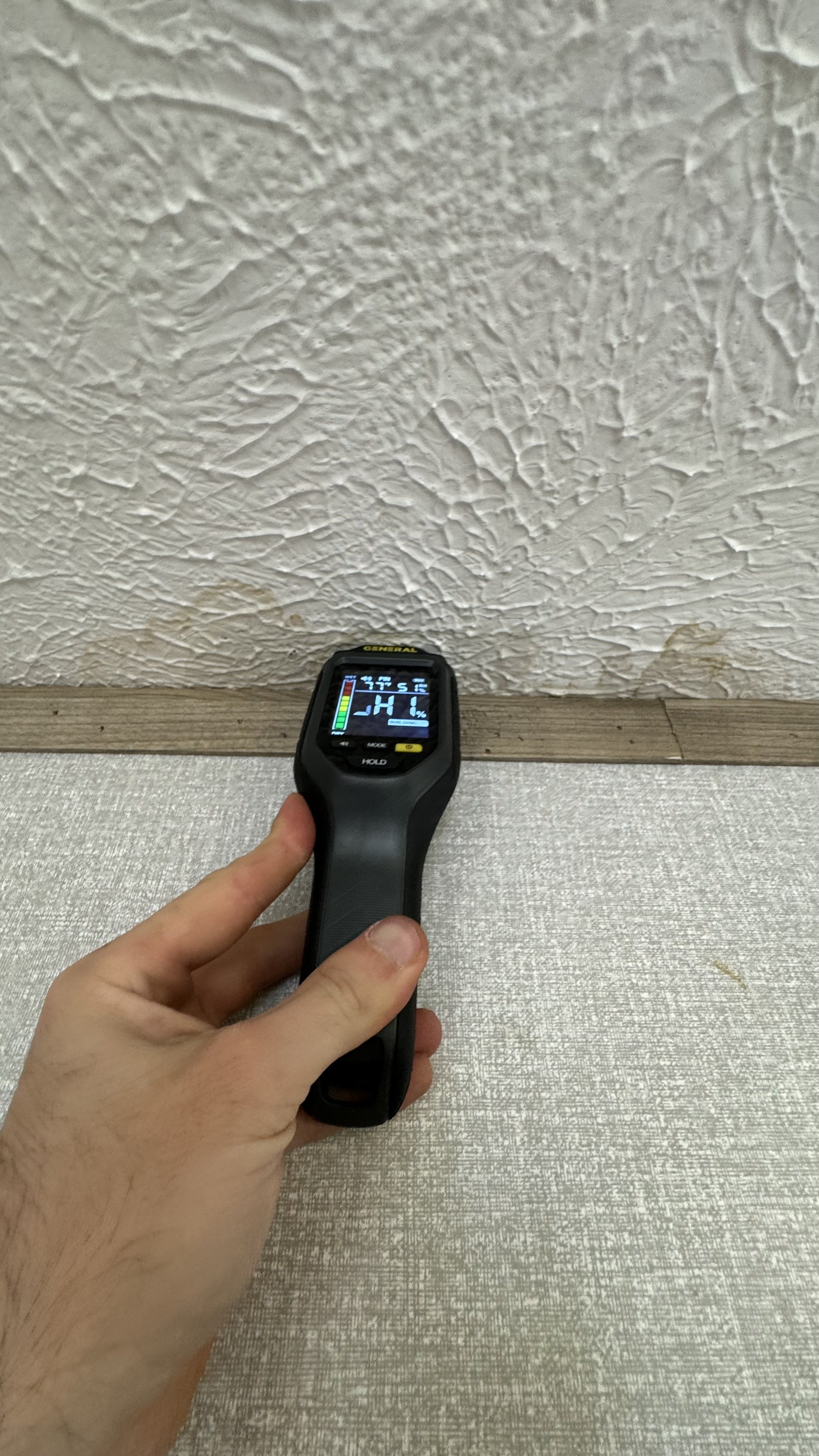 Person holding a digital moisture meter at a textured ceiling with high moisture intrusion.