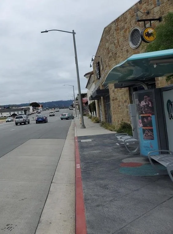 Empty sidewalk with benches outside a building with a yellow and black sign, overcast sky, and cars on the street. Urban environment in Monterey County, California.