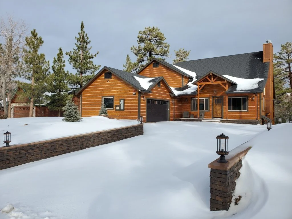A large wooden house with a black roof surrounded by snow and pine trees in Big Bear Lake, California.