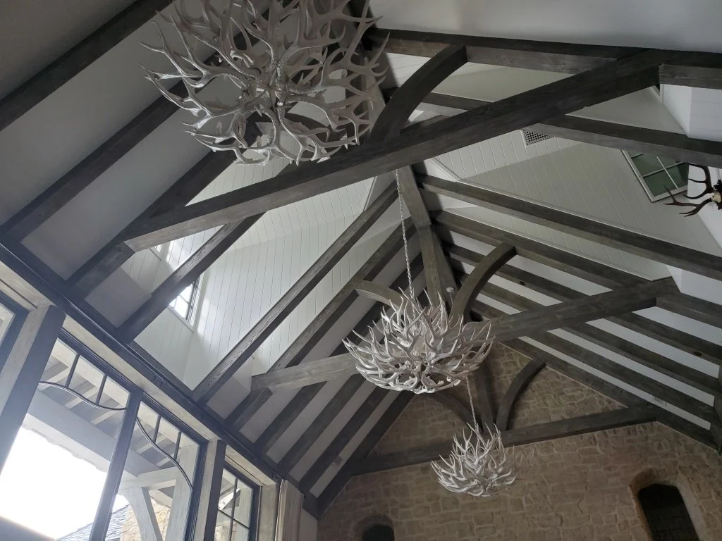 Interior view of a ceiling with exposed wooden beams and large decorative antler chandeliers hanging from it, with windows allowing natural light to enter.