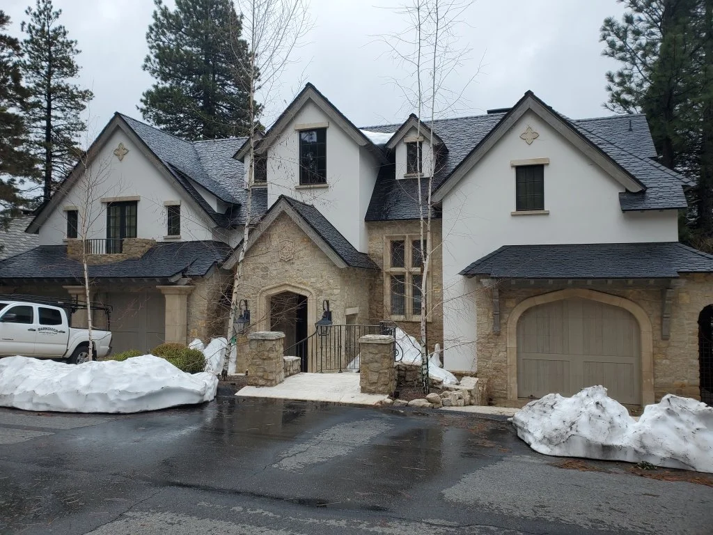 A large, multi-story house in Lake Arrowhead, CA, with a stone & white exterior, black roof, multiple windows, and an attached garage. There are patches of snow on the ground and wet pavement in front of the house, with a vehicle parked to the left.