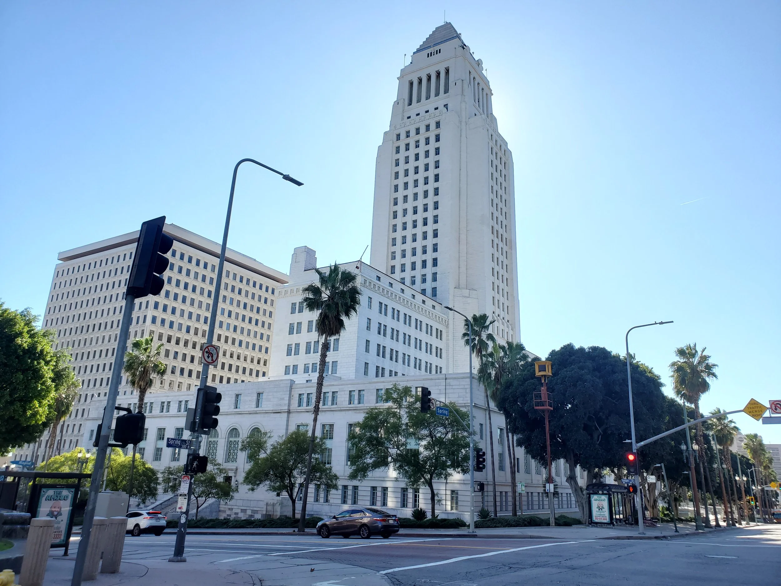 A city street scene featuring the Biltmore Hotel in Los Angeles, with tall buildings, palm trees, traffic lights, and cars at a red light.