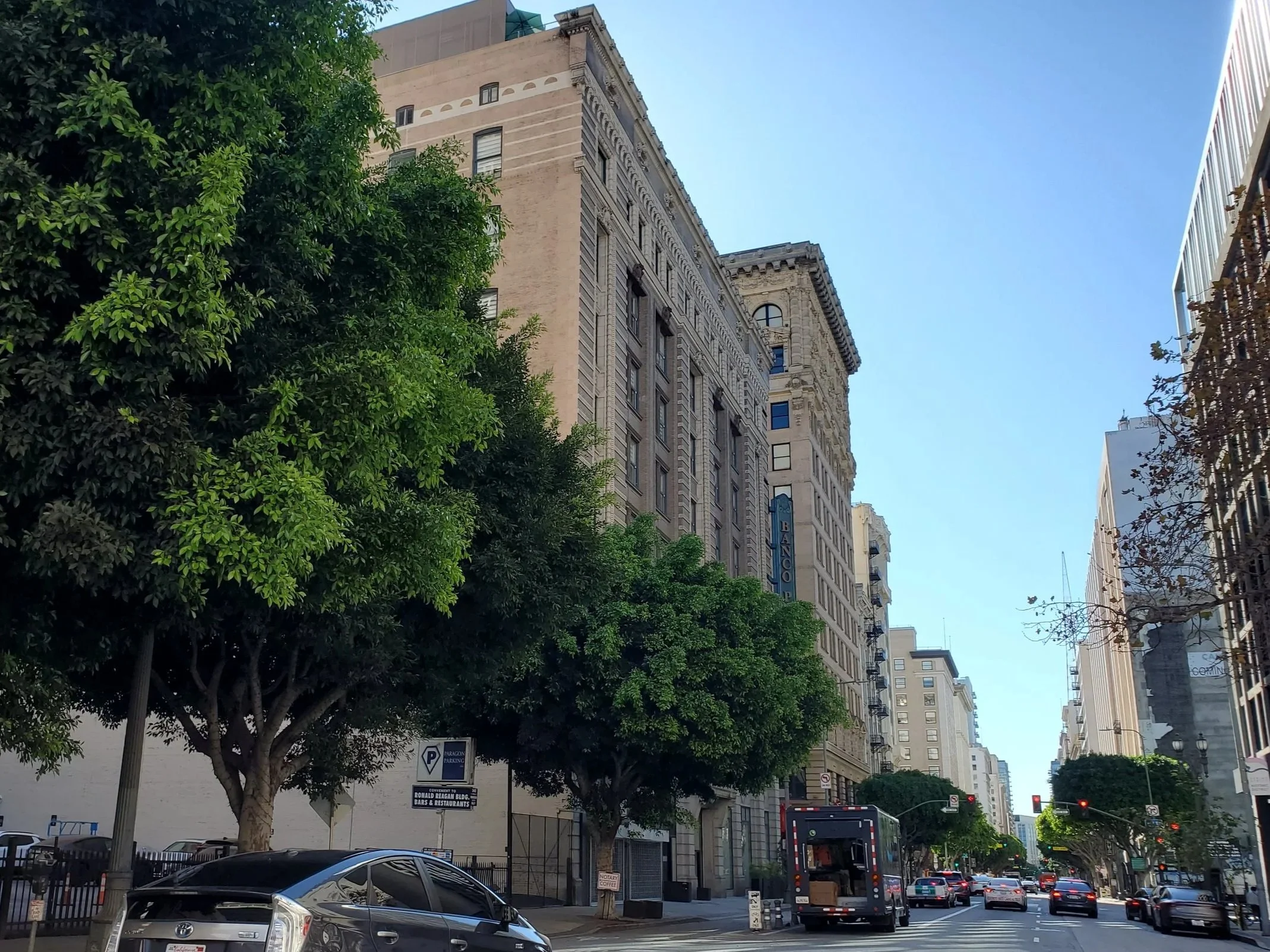Downtown Los Angeles street with tall buildings, trees lining the sidewalk, cars parked and driving, and traffic signals, under a clear blue sky.