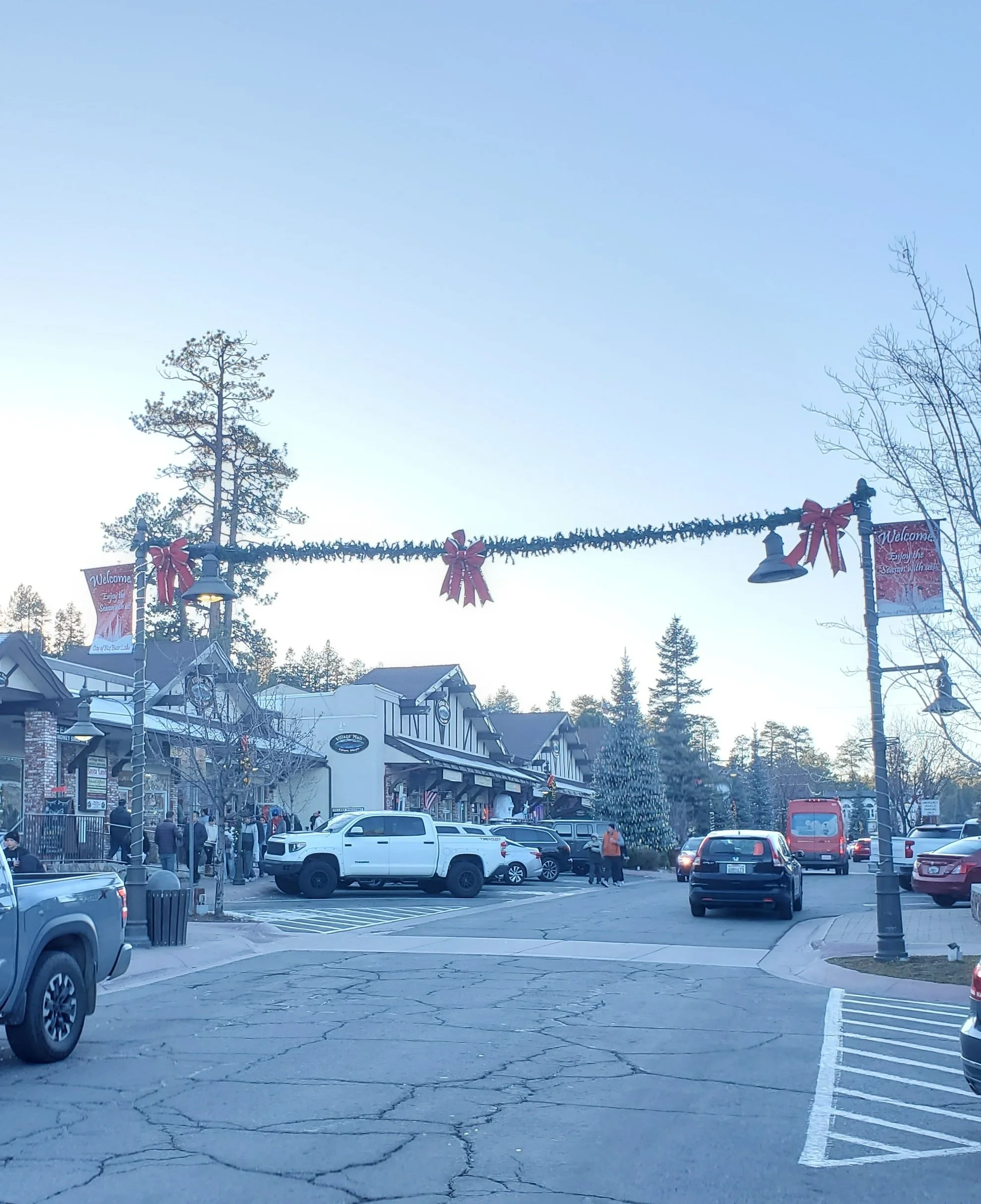 A small shopping plaza in Big Bear Lake Village, California, decorated with holiday decorations, including a string of lights and large red bows. There are several cars parked outside, and a few people walking near the shops.