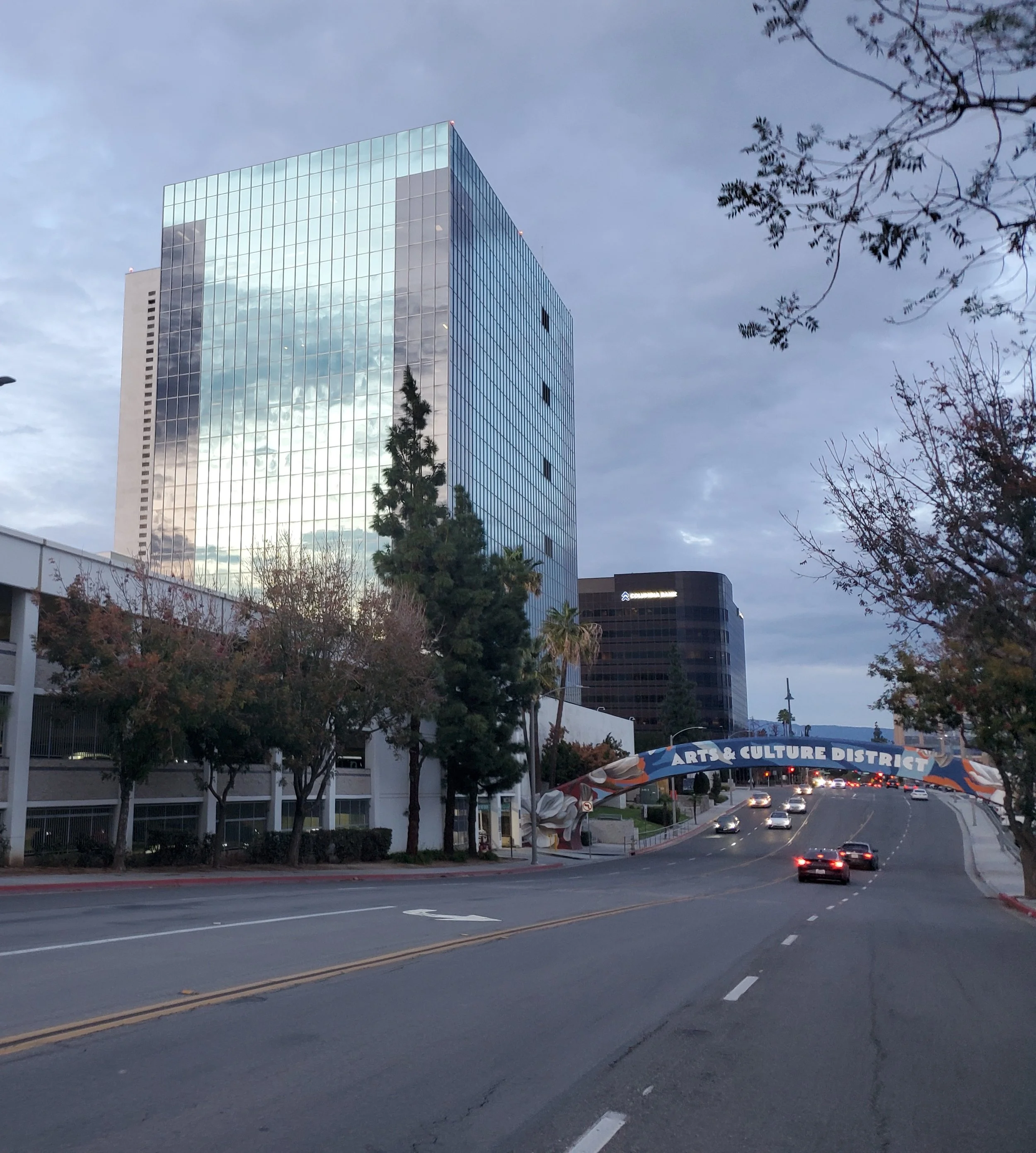 City street view in Riverside, California, with tall glass office buildings, trees on the sidewalks, and an arched sign that reads 'Arts & Culture District' over the road, with some cars driving.