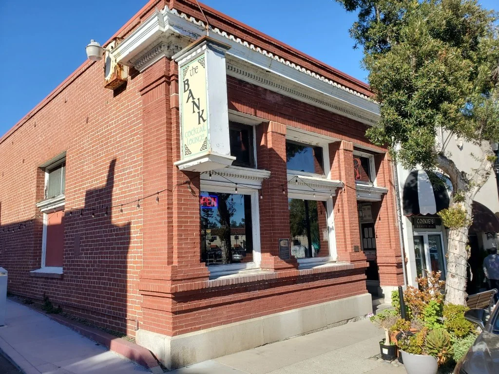 Brick building in downtown Saratoga, California, with a sign for 'The Bank' cocktail lounge, with an entrance door and windows, and a small shop nearby, with a tree and plants in front.