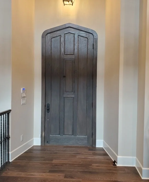 A dark wooden arched doorway on a beige wall, illuminated by a wall-mounted light. The floor is hardwood, and there is a small black railing on the left side.
