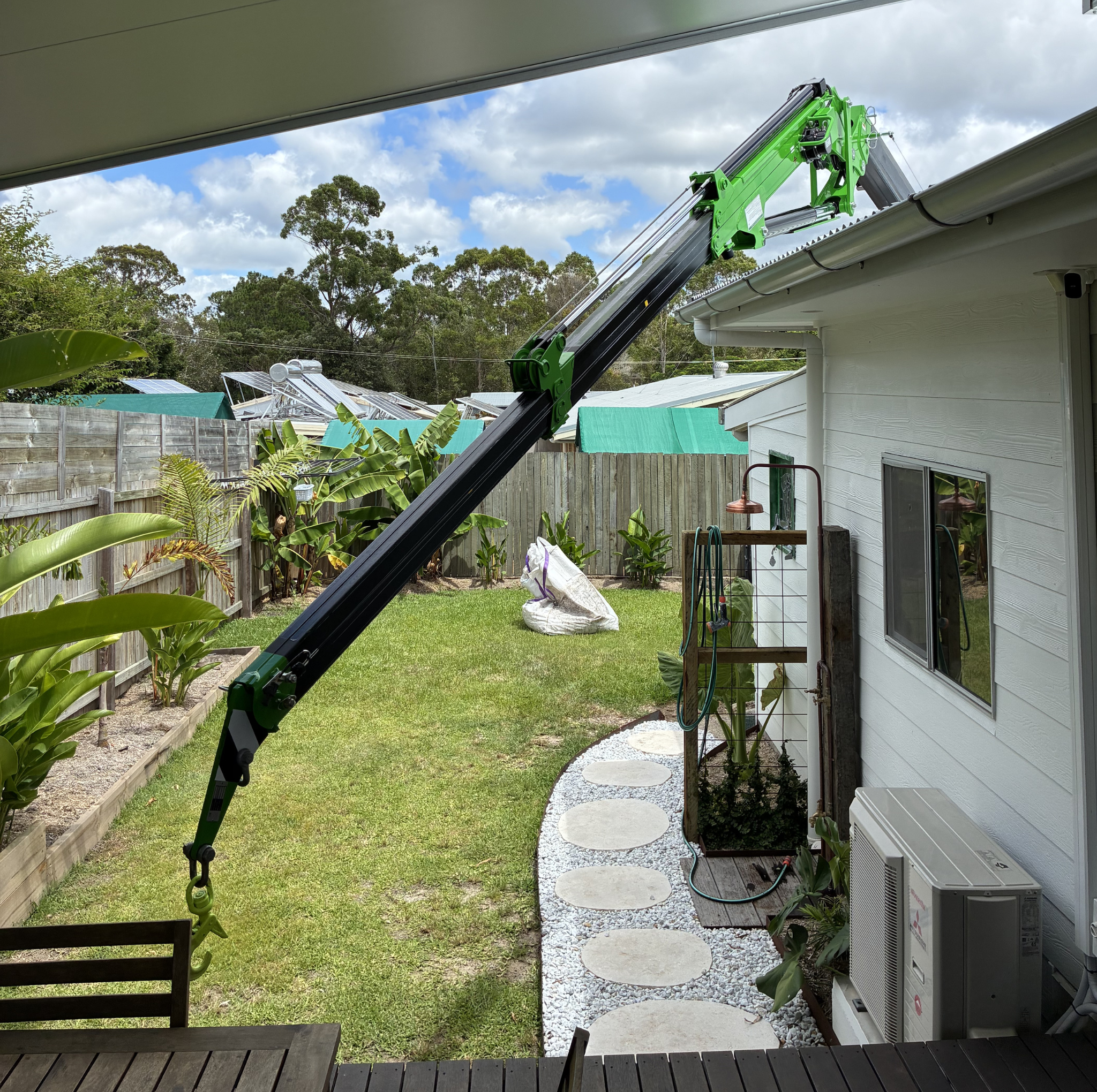Backyard with a white house, a wooden fence, green plants, a pathway with stepping stones, and a large black and green telescopic boom arm extending from a window on the house's wall.