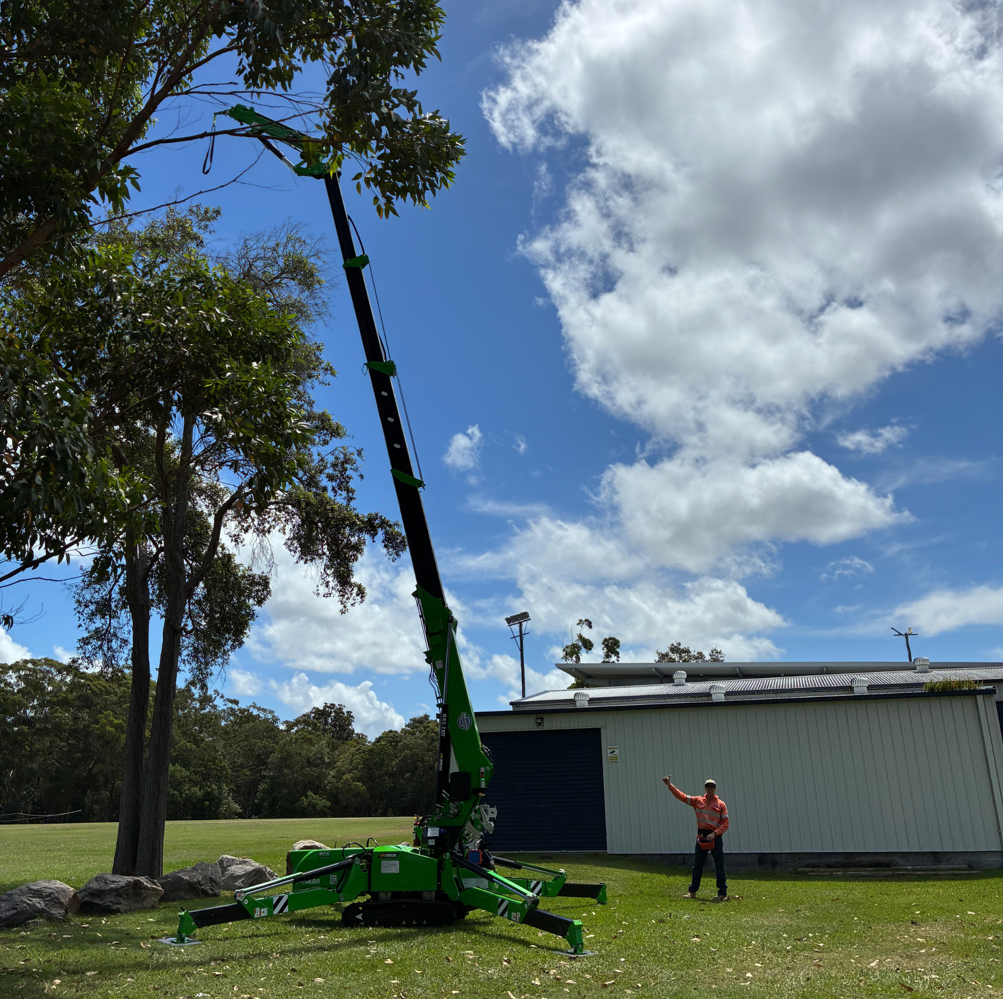 A man standing next to a green mini crane extended to reach a tree, with a white building and cloudy blue sky in the background.