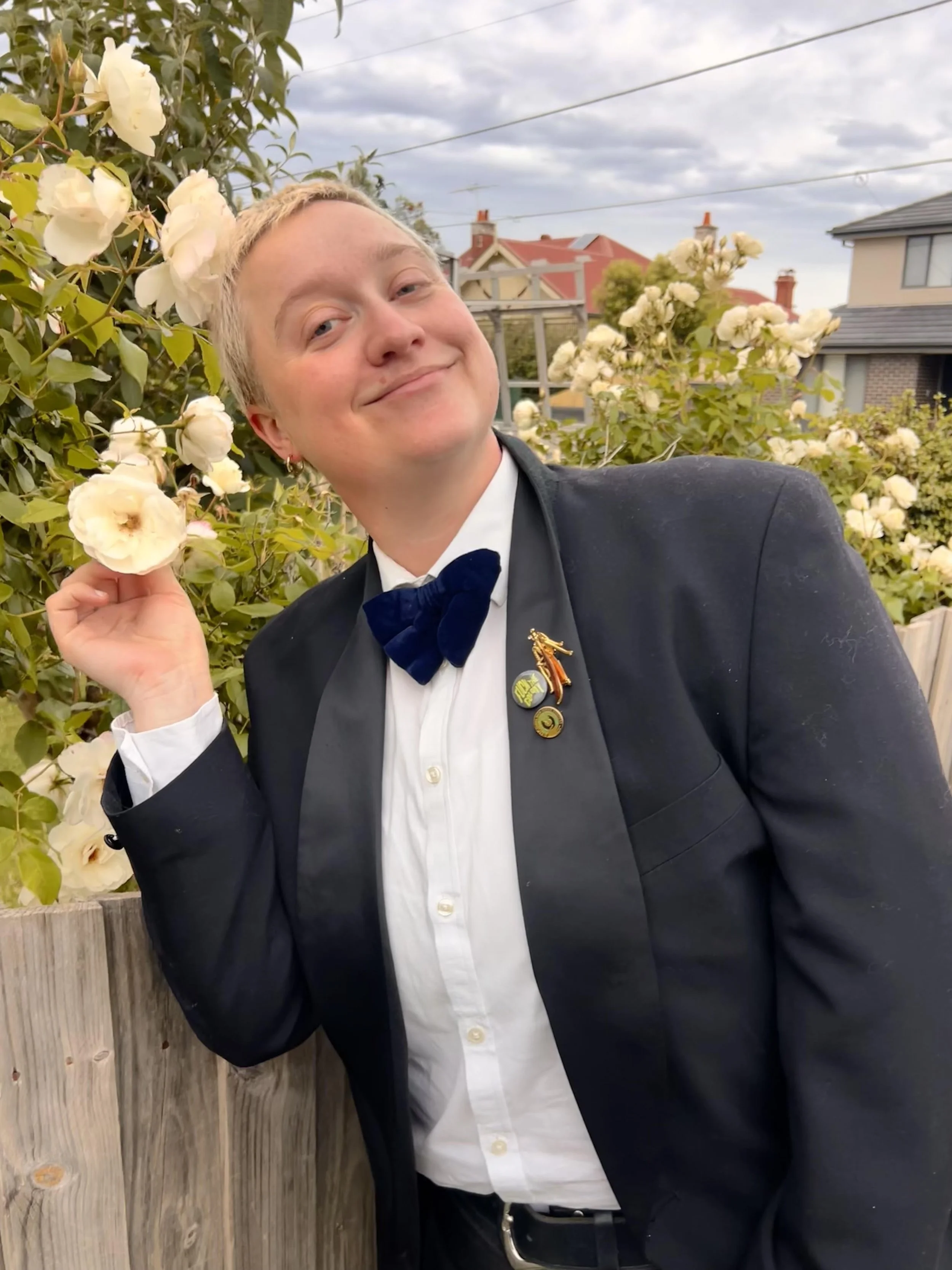 A person in a tuxedo with a bow tie, standing outdoors near a flower garden, smiling and holding a cream-colored flower with a background of houses and cloudy sky.