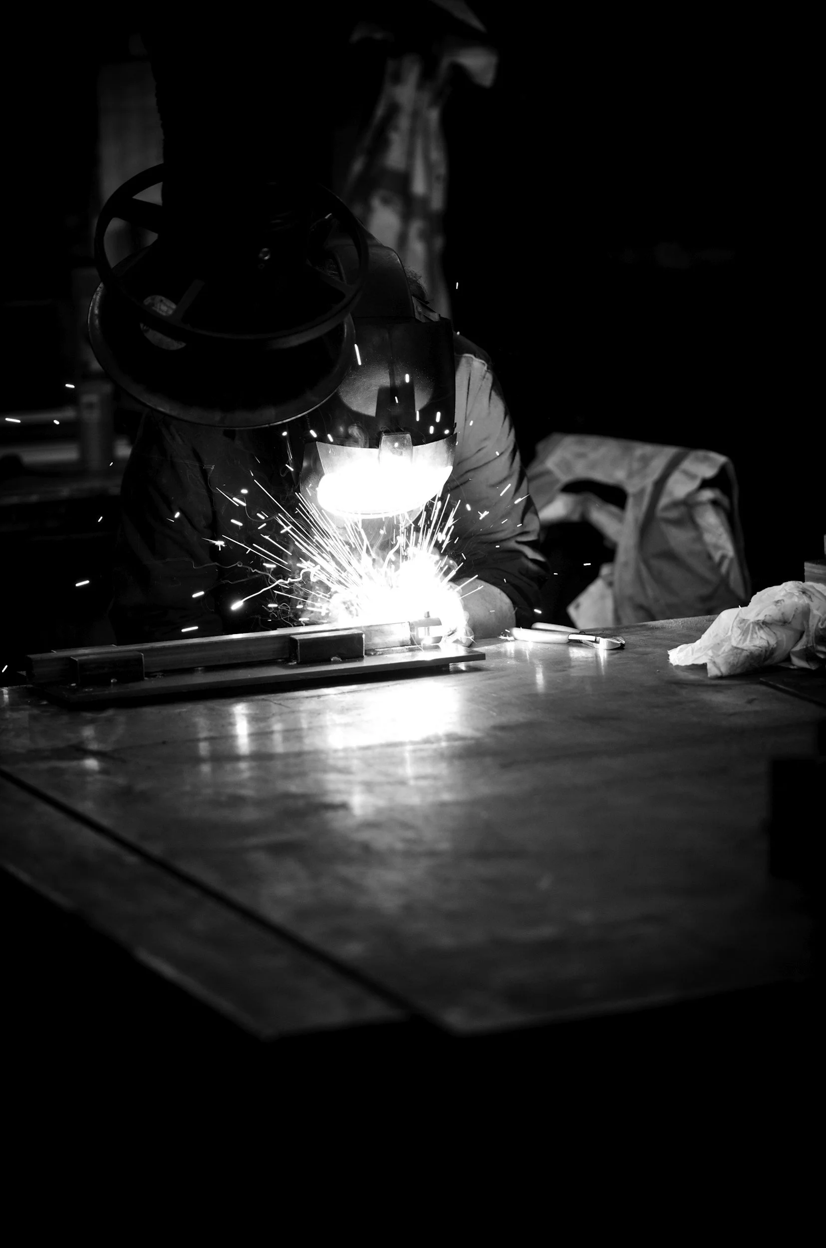 A person welding metal at a workbench, sparks flying, in a dark workshop.