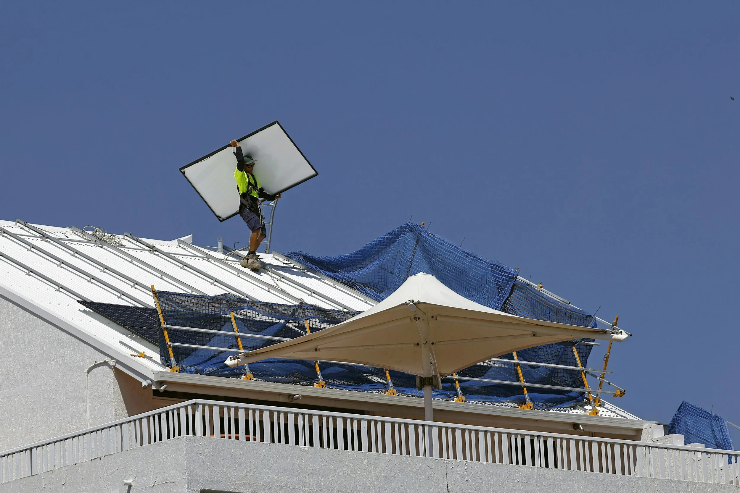 A construction worker in a green safety vest and dark helmet installing a solar panel on a white building's roof under a clear blue sky.