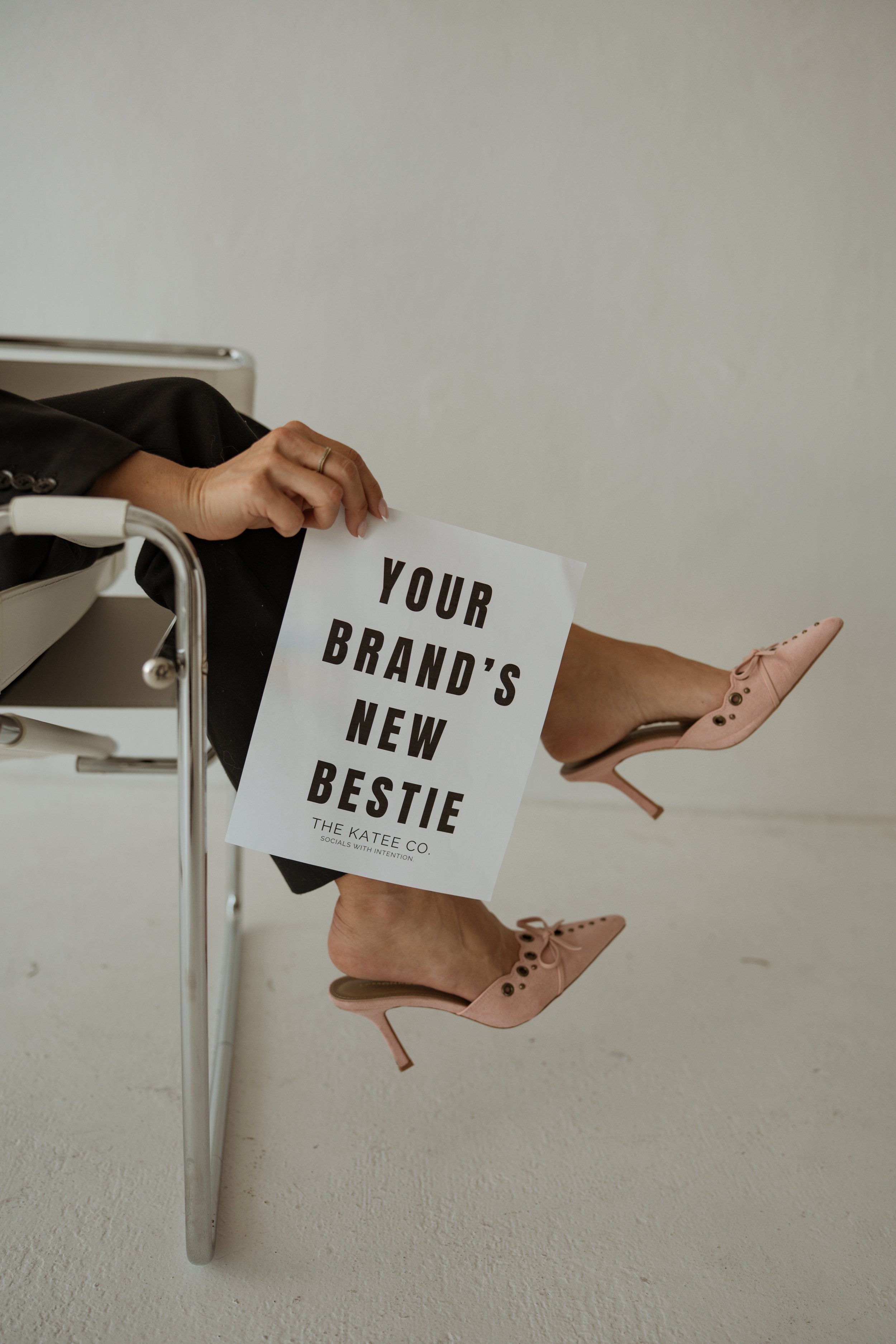 Person sitting on a chair with pink high heels, holding a sign that reads 'Your Brand's New Bestie' and 'The KATCEE Co.'
