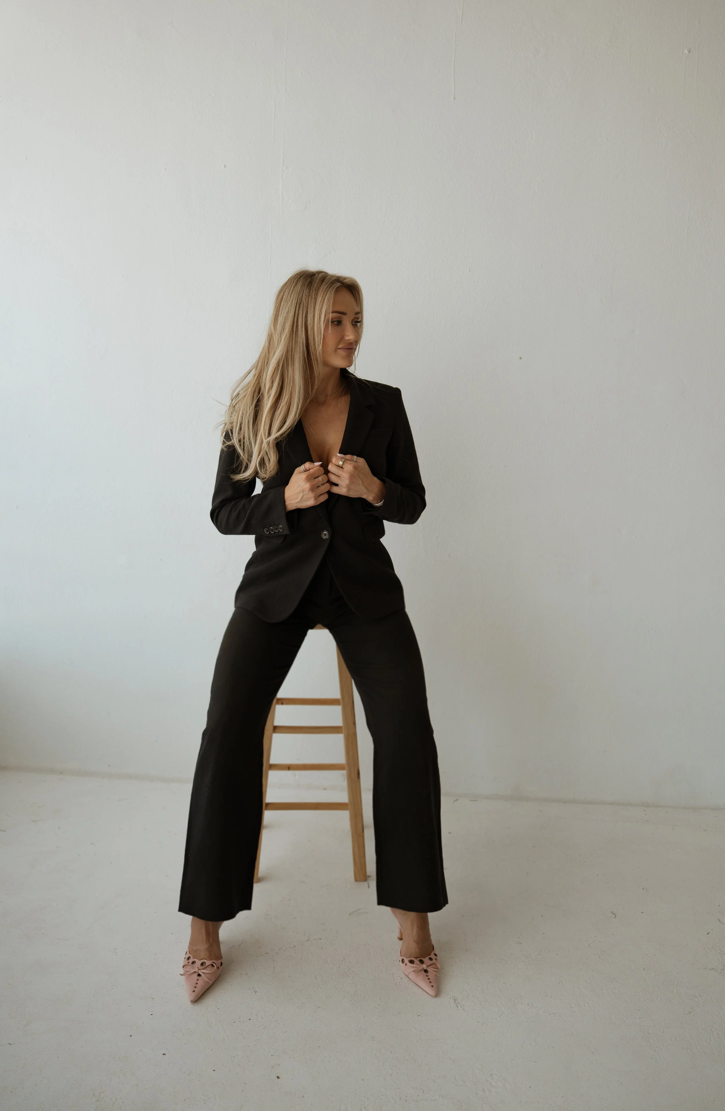 A woman with long blonde hair wearing a black suit and pink high heels, sitting on a wooden stool against a plain white wall.
