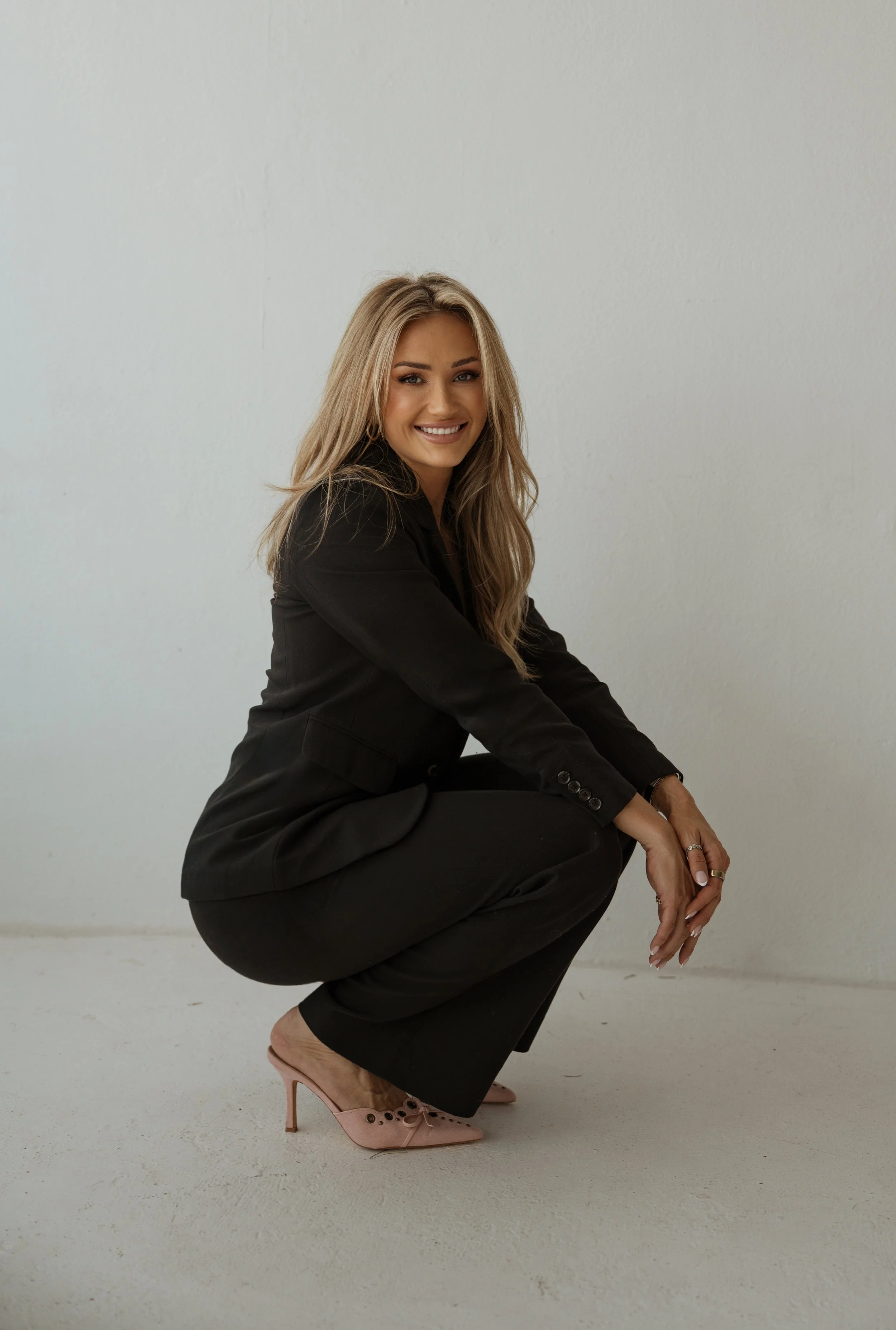 A woman with long blonde hair smiling and crouching on the floor, wearing a black suit and pink high heels, in a plain white room.