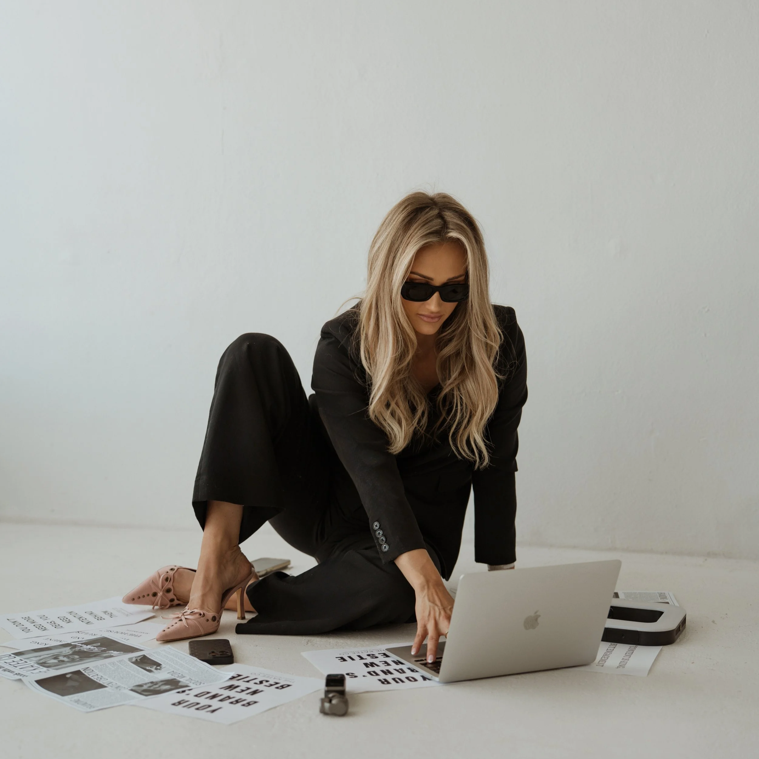 Woman in black suit with blonde hair, wearing sunglasses, sitting on the floor surrounded by newspapers, a laptop, and office equipment, in a white room.