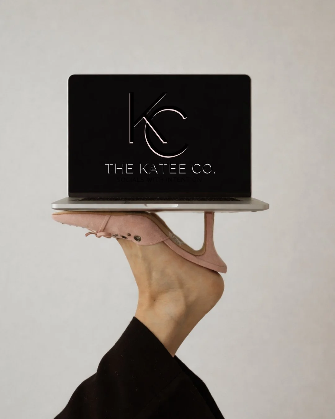Person holding a laptop with the logo 'KC' and the text 'The Katee Co. Socials with Intention' on the screen, wearing pink high heel shoes against a plain background.
