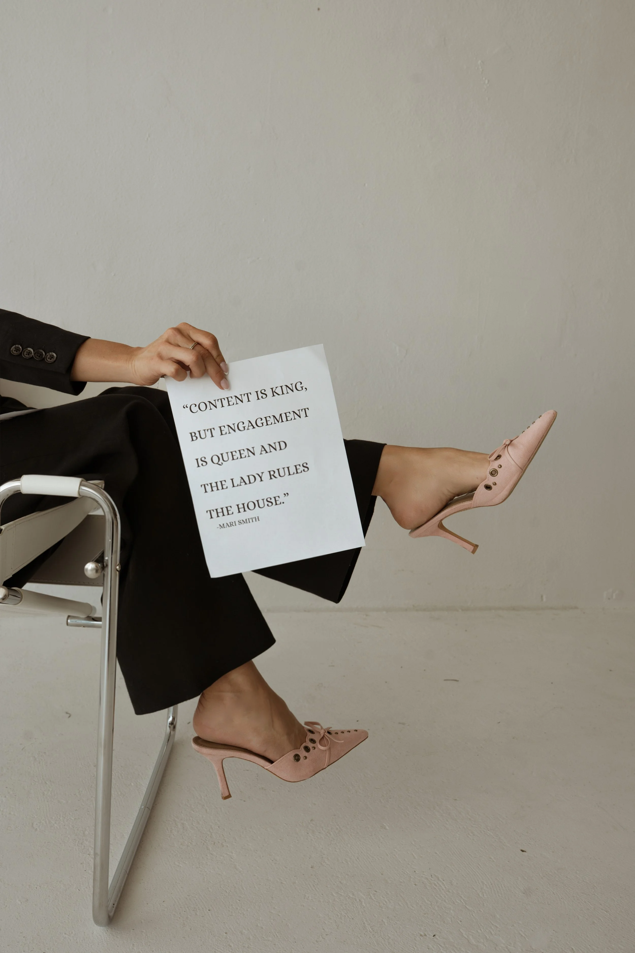 Person sitting in a chair with legs crossed, wearing black pants and pink high heels, holding a sign with the quote 'Content is king, but engagement is queen and the lady rules the house.'