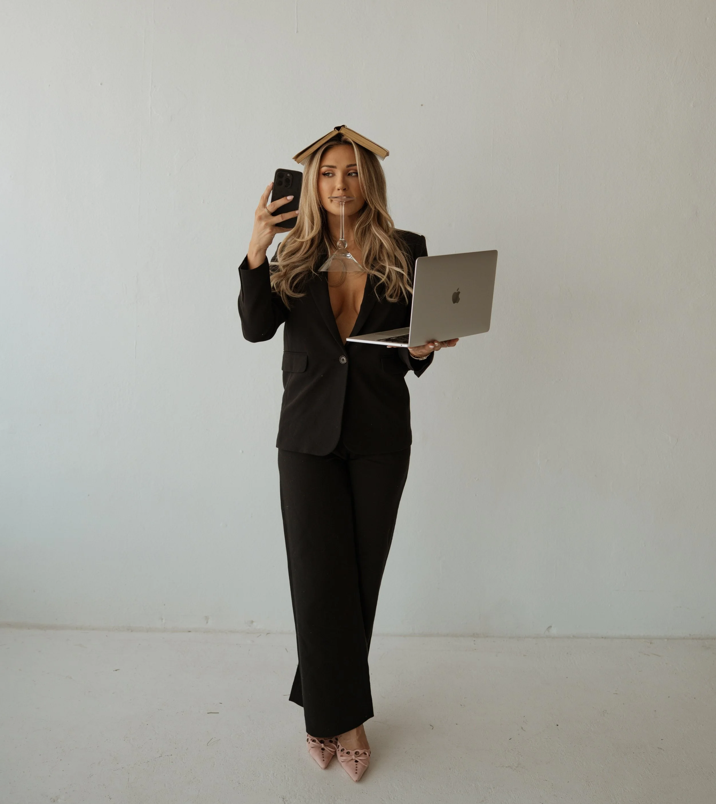Woman in black suit balancing a cup on her head, holding a laptop and a phone, against a plain wall.