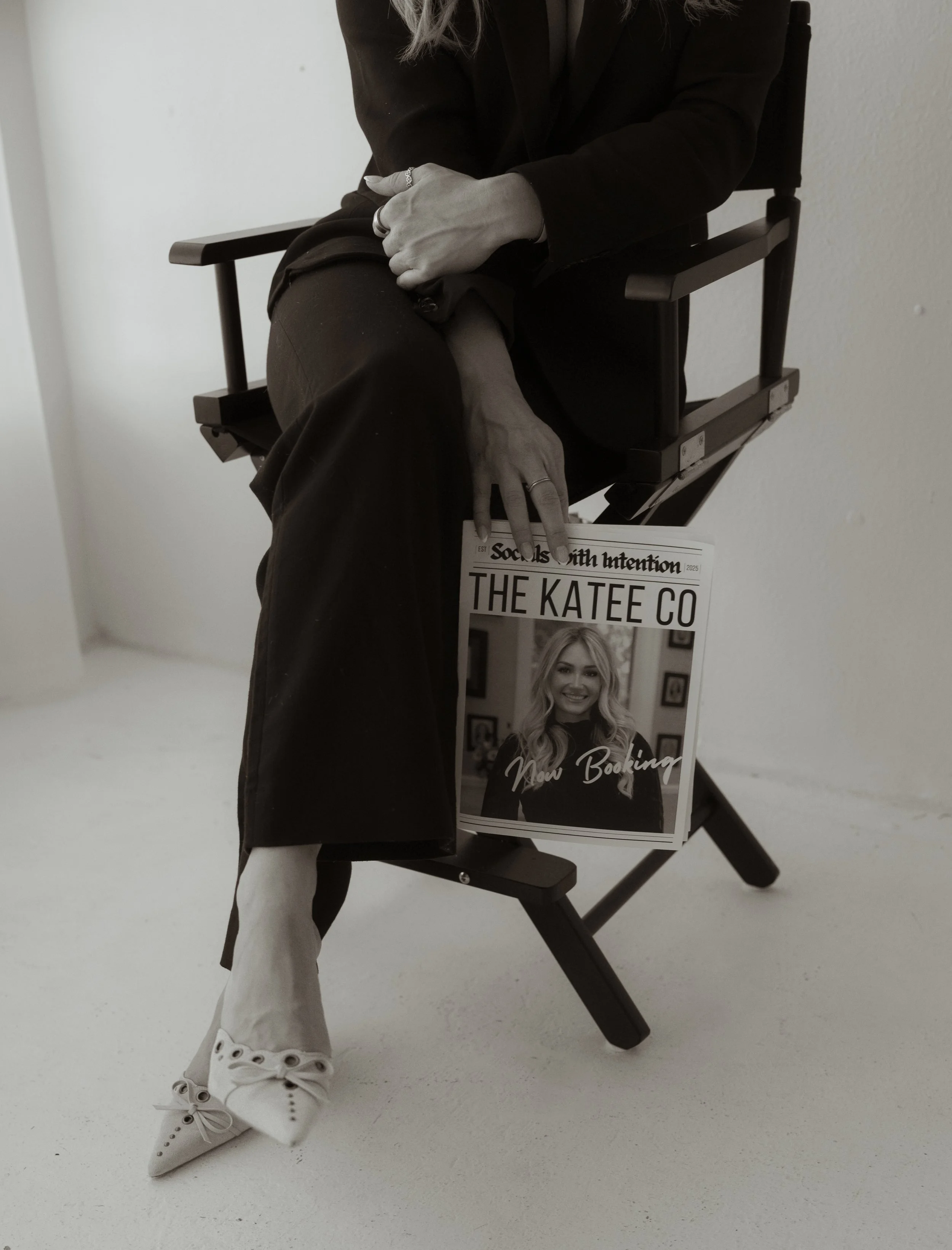 A woman sitting on a black director's chair, wearing a black outfit, with her hands resting on her lap and a magazine titled 'The Katee Co' hanging from the chair's side. The magazine features a smiling woman on the cover.