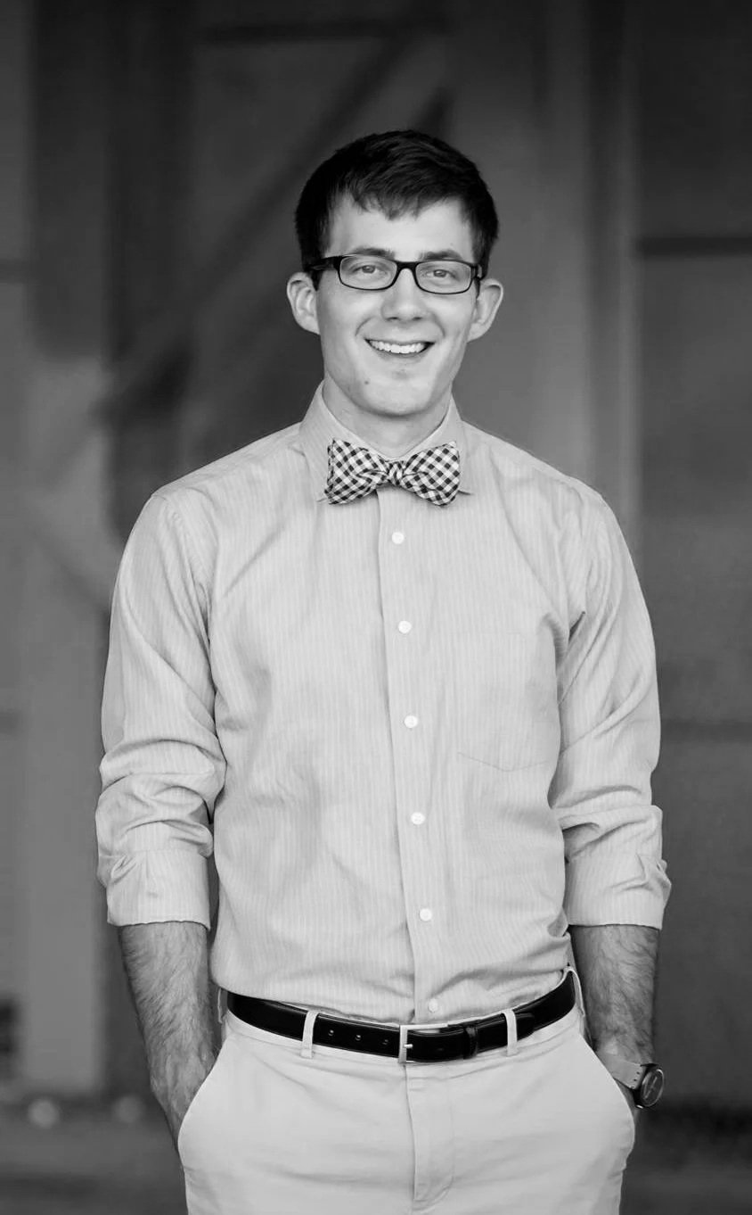 A young man with glasses, wearing a checkered bow tie, a button-up shirt, and light-colored pants, smiling with hands in pockets, in a professional setting.