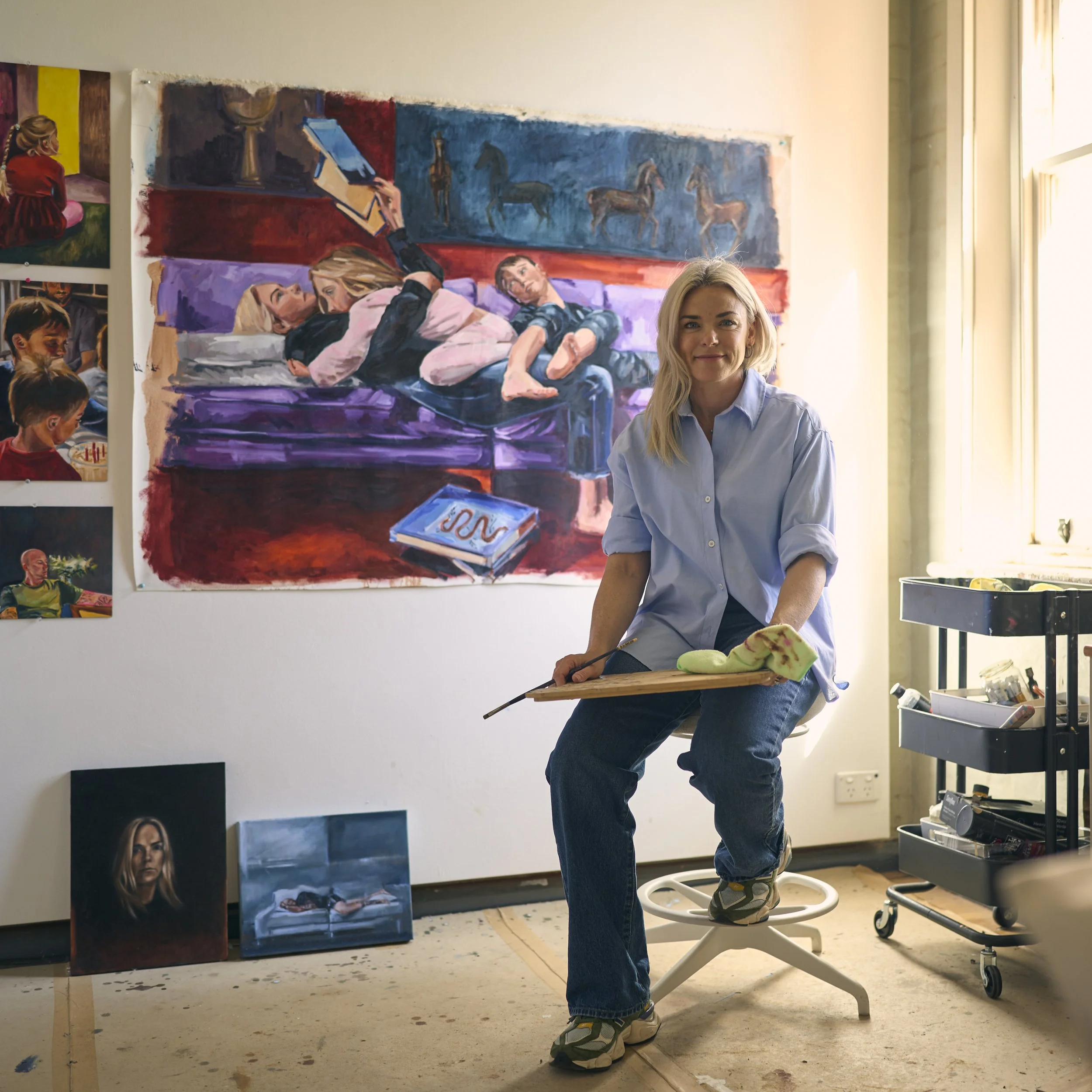 A woman artist sitting on a stool in her studio, holding a paint palette, with a large colorful painting of children on a bed hanging on the wall behind her. Various artworks and art supplies are visible around her.