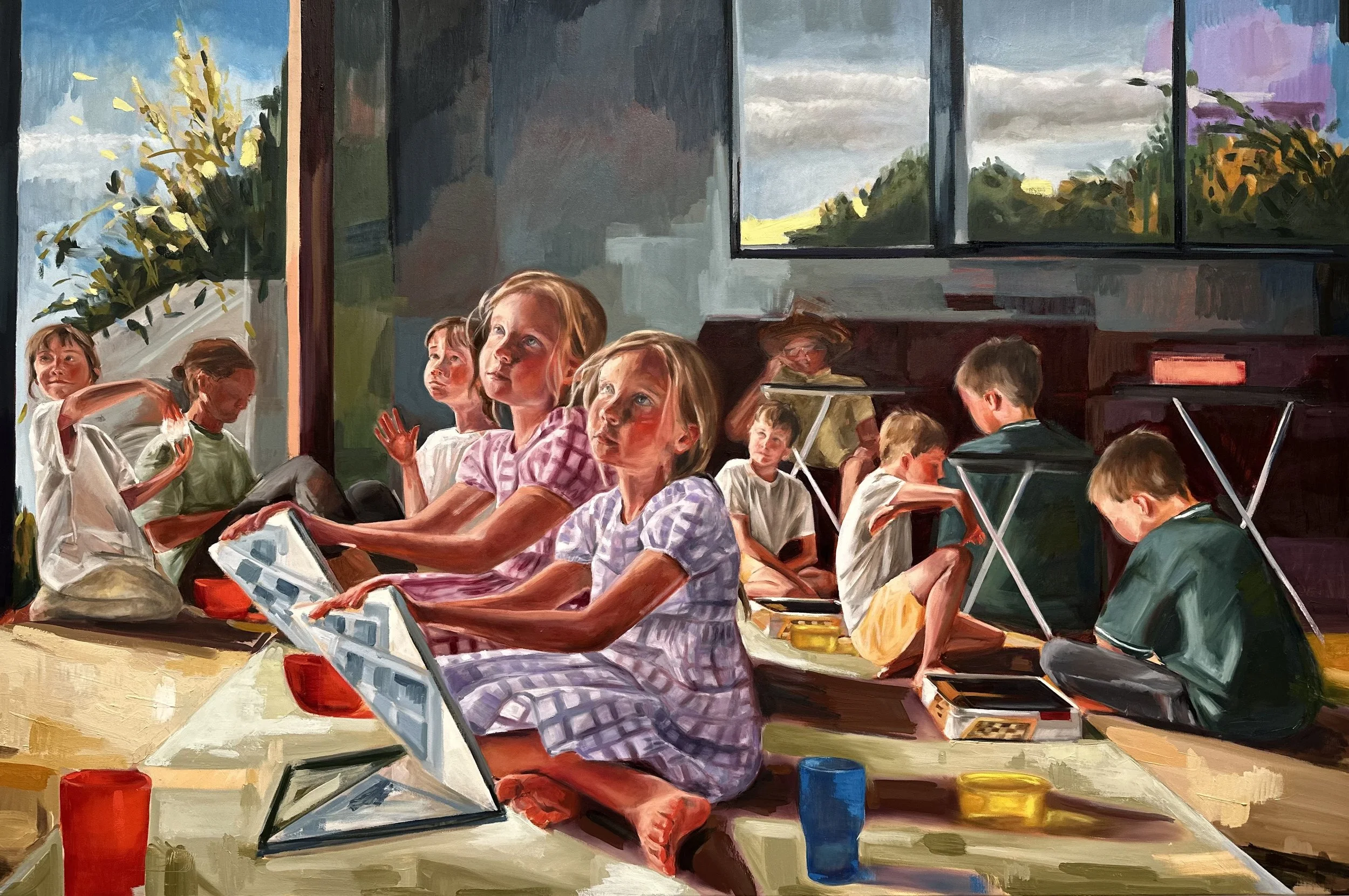 Children sitting on the floor in a classroom, engaging in activities. Some are playing and others are watching. Large windows with a view of trees and the sky in the background.