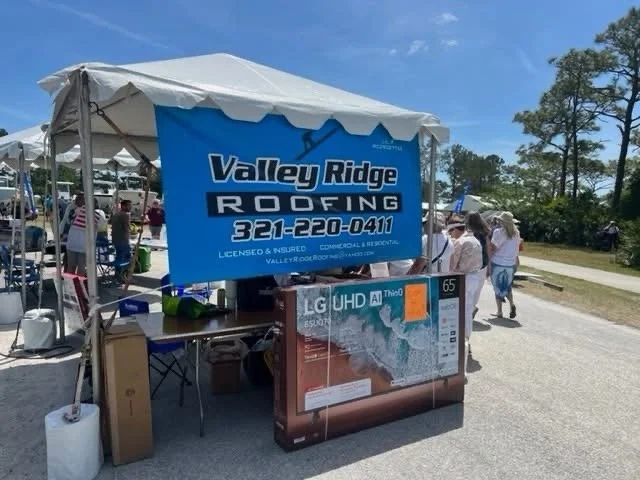 Outdoor market tent with a blue Valley Ridge Roofing sign, people walking nearby, and a large LG UHD TV on display.