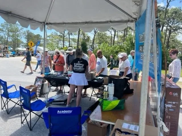 People under a tent at an outdoor event, participating in a cooking or food preparation activity, with tables and chairs around.