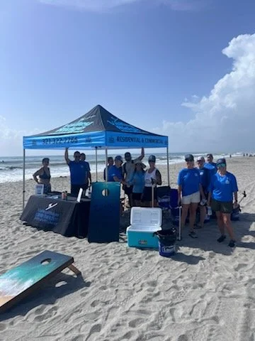 People under a blue tent on the beach, with equipment on tables and sand in the background.