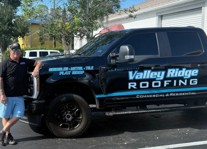 A man standing next to a black pickup truck with blue and white Valley Ridge Roofing branding, parked in a driveway with trees and houses in the background.