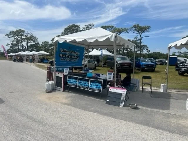 Outdoor tent booth for Valley Ridge Roofing, with promotional signs, chairs, and a menu, set up along a gravel road with parked cars and trees in the background.