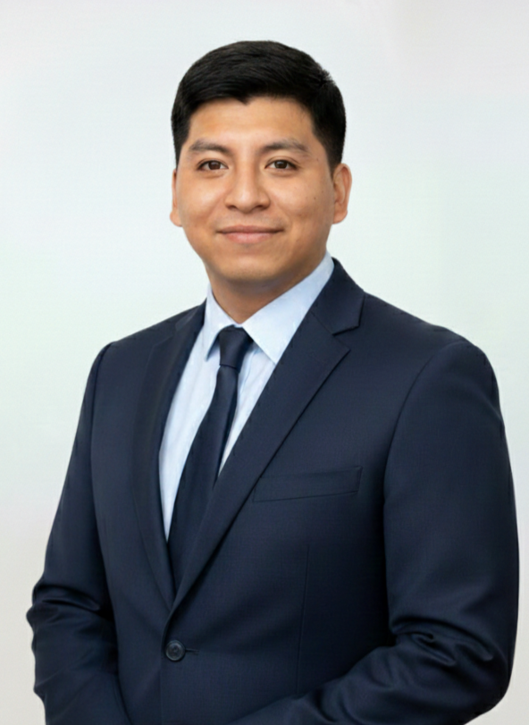 A professional man wearing a dark suit, light blue shirt, and tie, standing against a plain background, smiling at the camera.
